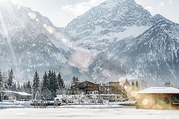 Hotel am See vor verschneiten Bergen mit Sonnenlicht und winterlicher Stimmung