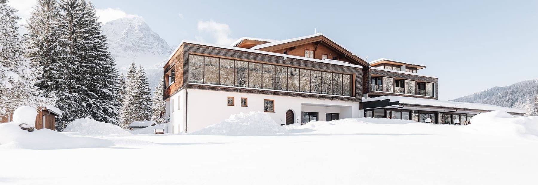 Zeitgenössisches Hotelgebäude mit großen Fensterflächen in verschneiter Berglandschaft bei klarem Himmel.