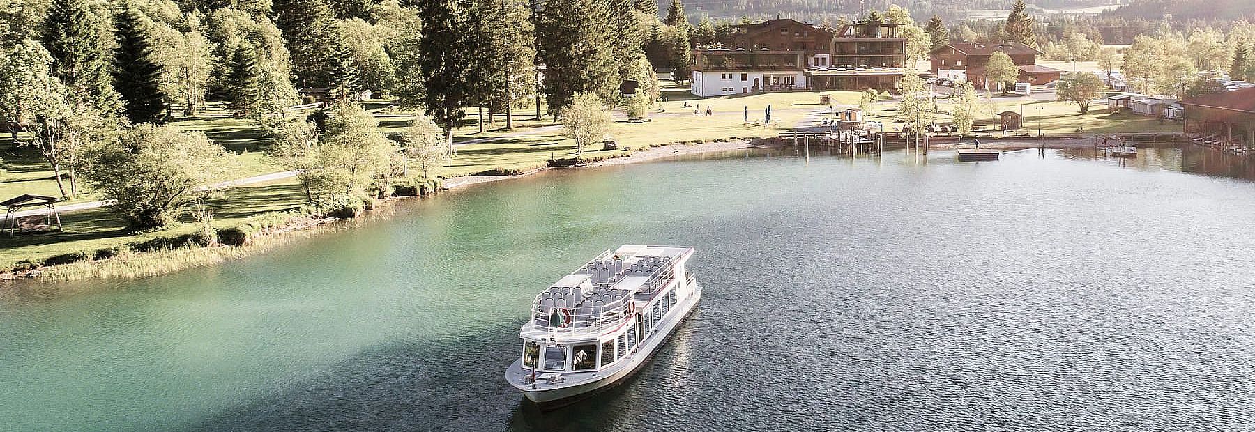Ausflugsschiff vor dem Ufer mit Wald, Wiese und Hotelanlage bei Sonnenschein