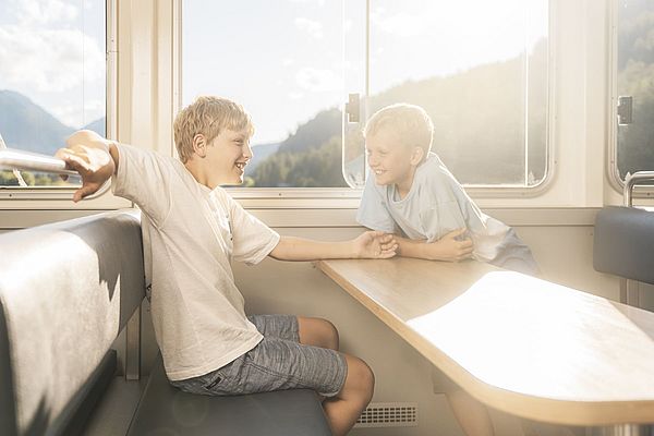 Zwei Jungen sitzen lachend am Tisch im Schiffsinneren mit Sonnenlicht und Bergblick