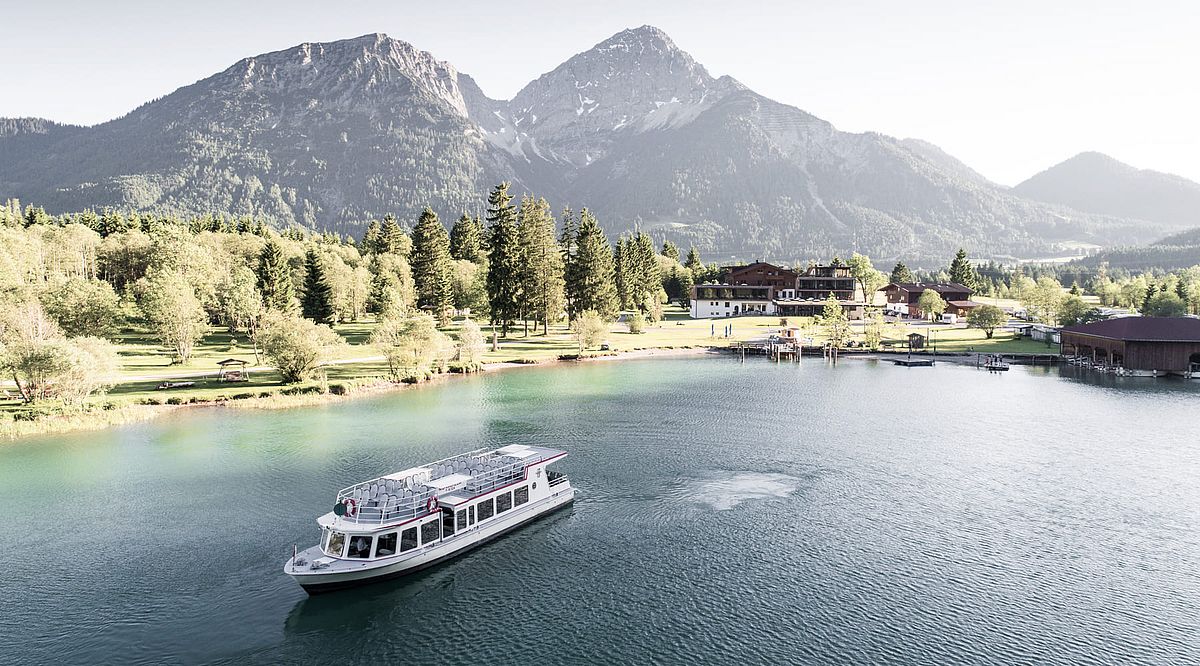 Ausflugsschiff auf türkisfarbenem See mit Bergen, Wald und Hotel im Hintergrund