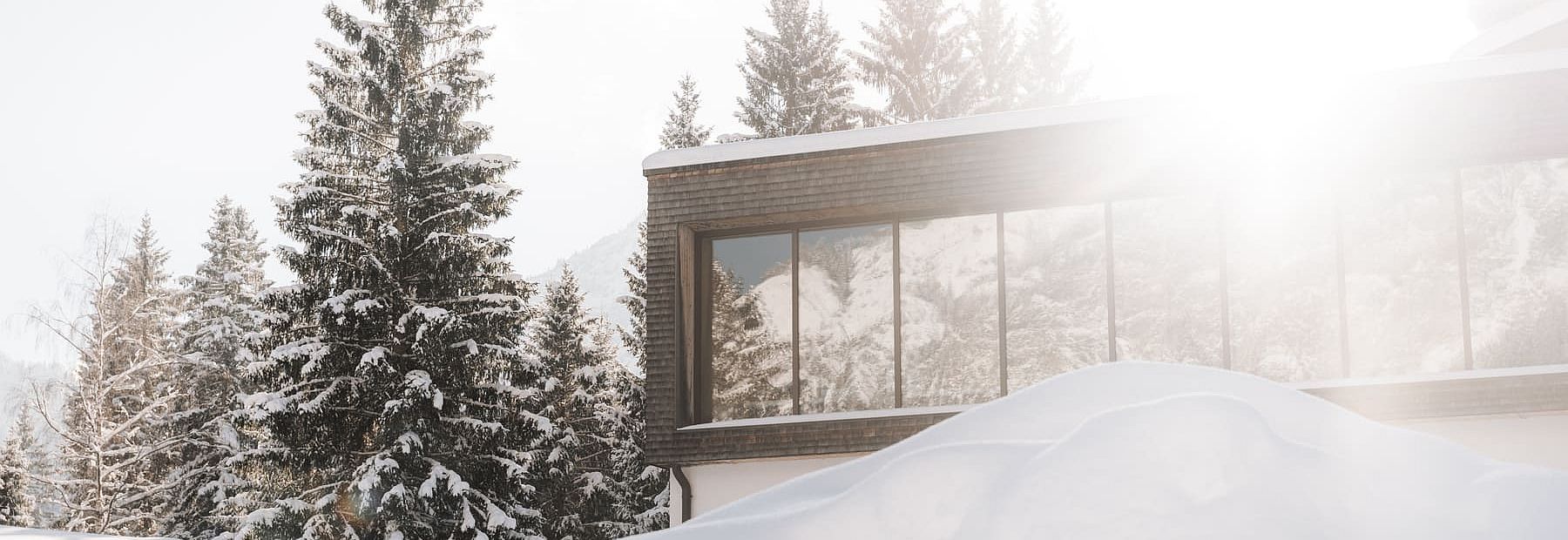 Verschneite Winterlandschaft mit Blick auf eine moderne Hotelfassade, Sonnenlicht über Tannenbäumen.
