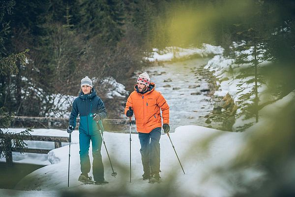 Zwei Schneeschuhwanderer unterwegs im Winterwald an einem verschneiten Bachlauf
