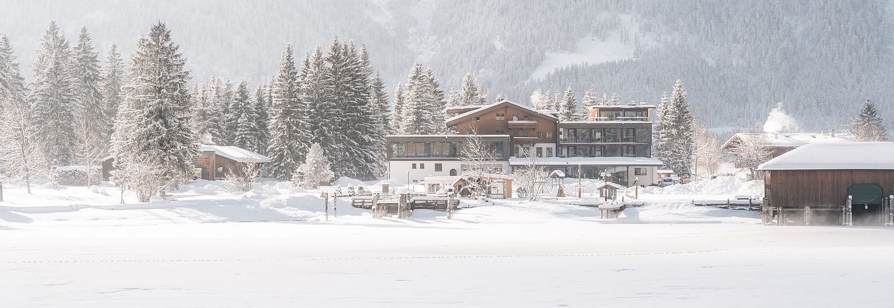 Modernes alpines Hotelensemble im Winter, umgeben von verschneiten Tannen und Bergen, Blick über eine schneebedeckte Fläche.