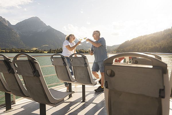 Paar stößt mit Weingläsern auf dem Sonnendeck eines Schiffs mit Bergkulisse an